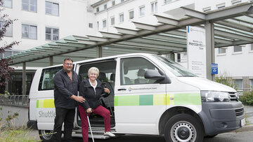 Fahrer des Patientenshuttle hilft Patientin mit Krücken in den Kleinbus.