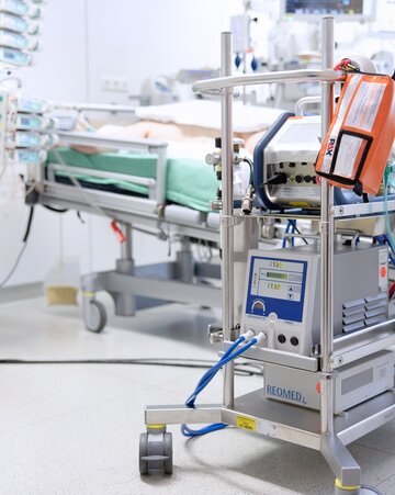 Moderne medizinische Geräte mit einem orangefarbenen Defibrillator und verschiedenen Monitoren auf einem Metallwagen in einem Krankenhauszimmer, die eine Atmosphäre klinischer Präzision vermitteln.